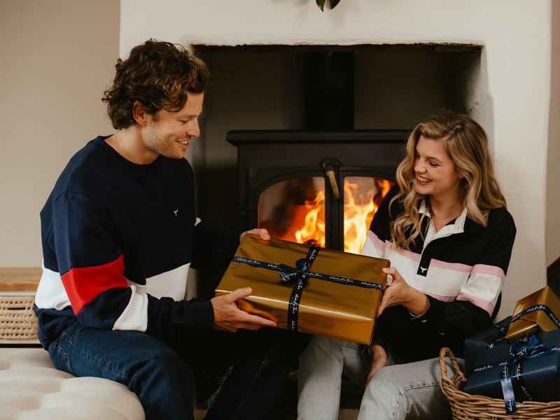 Man and woman exchanging a gold wrapped present in front of a fire 